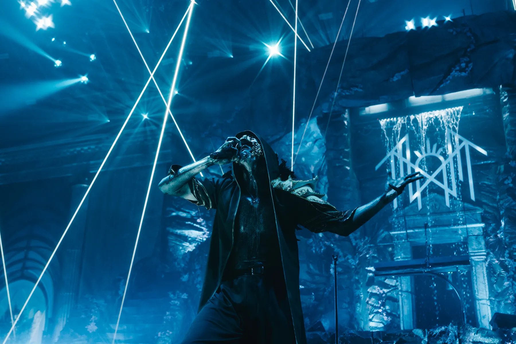 Masked, hooded performer on stage under laser beams and blue light